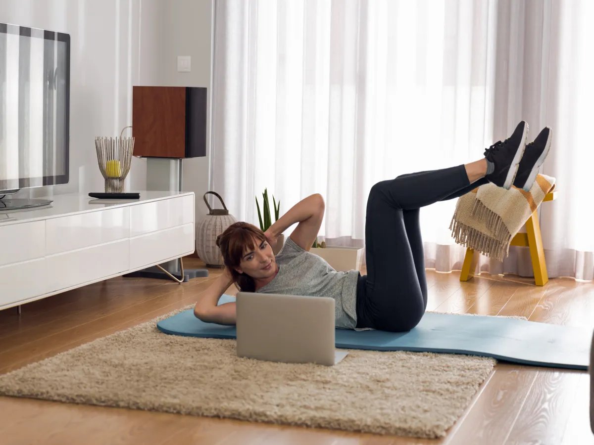 Determined to stay fit, a woman practices her workout routine at home, following an online class on her laptop. The warm and inviting living room sets the perfect backdrop for her dedication to health and wellness, surrounded by stylish decor and natural light streaming through the windows.