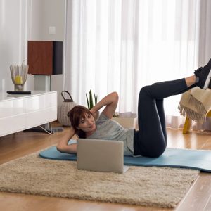 Determined to stay fit, a woman practices her workout routine at home, following an online class on her laptop. The warm and inviting living room sets the perfect backdrop for her dedication to health and wellness, surrounded by stylish decor and natural light streaming through the windows.
