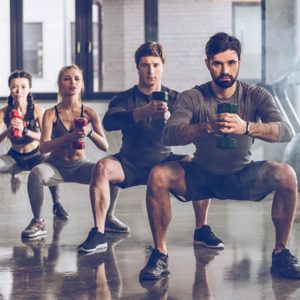 In a modern gym setting, four fitness enthusiasts engage in a synchronized squat exercise, emphasizing strength and teamwork while holding their weights. The atmosphere is charged with motivation and determination, as they strive for their fitness goals together.
