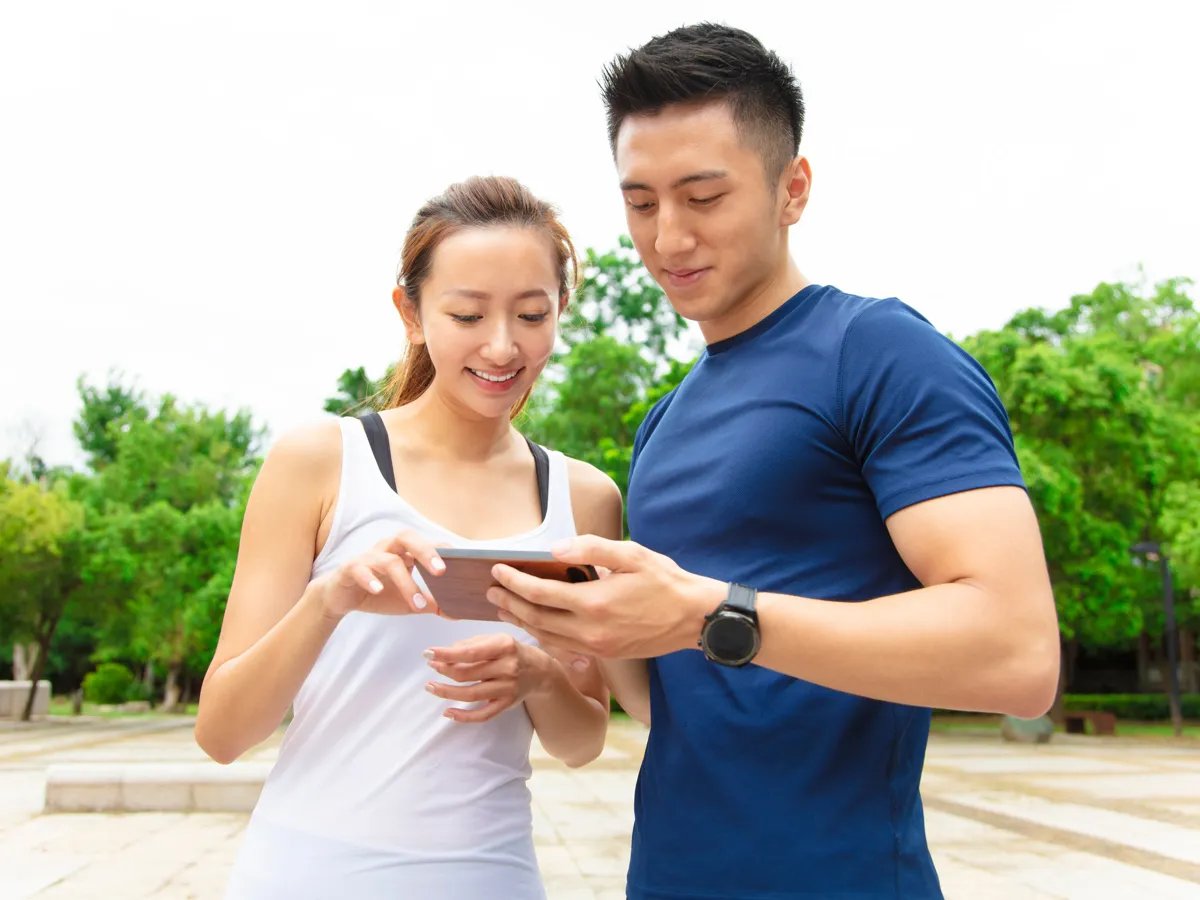 A young couple enjoys quality time outdoors, sharing a moment of laughter as they explore an app on their smartphone, surrounded by lush green trees and a scenic park setting.