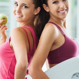 Two fitness enthusiasts stand back to back, exuding confidence in their health journey. One holds a fresh green apple, symbolizing nutritious choices, while the other proudly displays a scale, representing their commitment to fitness and well-being. Together, they embody strength and motivation in a vibrant workout environment.