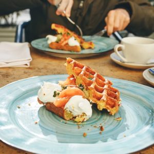 A delightful brunch scene unfolds, featuring a beautifully plated waffle topped with creamy yogurt and fresh herbs, while a diner savors each bite. The warm atmosphere invites relaxation and enjoyment, as steaming cups of coffee await at the table.