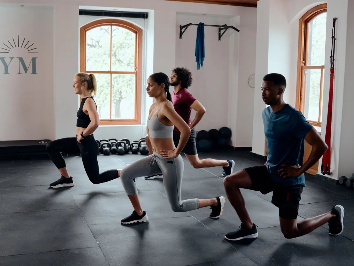 A dynamic group workout session unfolds in a well-lit gym, showcasing individuals dedicated to their fitness goals. With an assortment of exercise gear in the background, the participants execute lunging exercises, embodying strength and determination.
