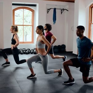 A dynamic group workout session unfolds in a well-lit gym, showcasing individuals dedicated to their fitness goals. With an assortment of exercise gear in the background, the participants execute lunging exercises, embodying strength and determination.