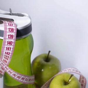 A refreshing blend of health and vitality, this vibrant green smoothie sits beside crisp green apples, promoting a wholesome lifestyle. A measuring tape wraps around the bottle, symbolizing fitness goals and the journey to well-being.