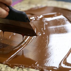 A skilled chocolatier glides a spatula over a luscious layer of melted chocolate, expertly tempering it on a cool marble surface. The rich, glossy texture promises a delightful treat that will soon be crafted into artisanal chocolate delights.