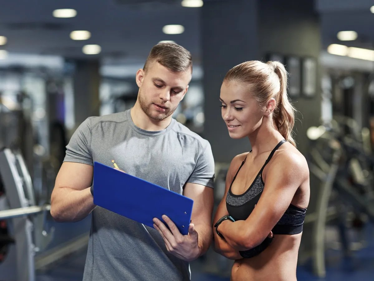 In the heart of a bustling gym, a dedicated trainer reviews workout plans with a motivated athlete, both focused on achieving their fitness goals together.