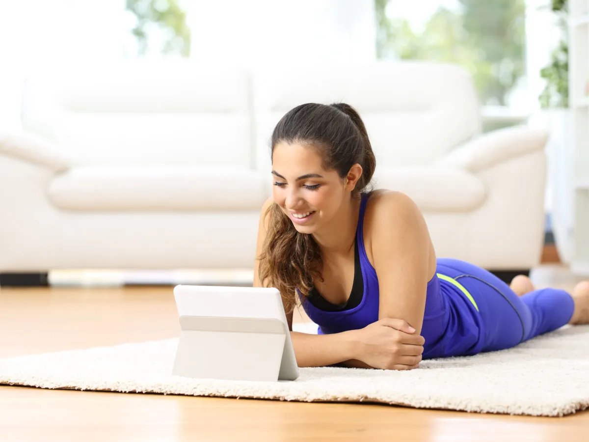 A young woman enjoys her leisure time, lying comfortably on a soft rug while browsing on her tablet. The bright, modern living room around her creates a welcoming atmosphere, perfect for relaxation and creativity.