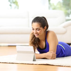 A young woman enjoys her leisure time, lying comfortably on a soft rug while browsing on her tablet. The bright, modern living room around her creates a welcoming atmosphere, perfect for relaxation and creativity.