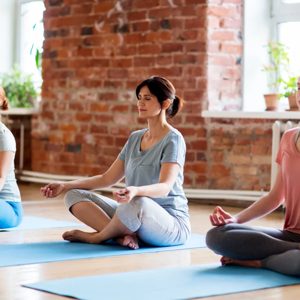 In a serene studio filled with natural light, three women practice meditation, finding their inner peace on yoga mats. The warm brick walls and the calming atmosphere set the perfect scene for reflection and mindfulness.